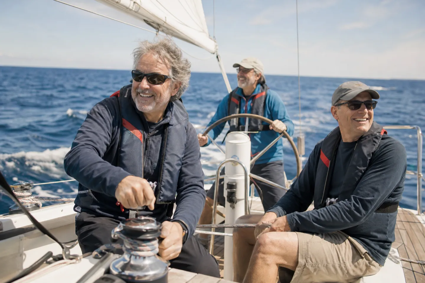 A skipper trims a sheet on a winch while two crew smile at the helm, offshore